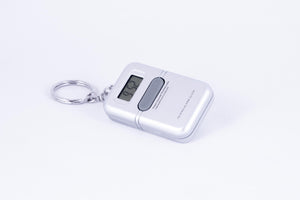 A silver, rectangular, hand-held digital clock on a keychain, with one central button to request audible time in Spanish.